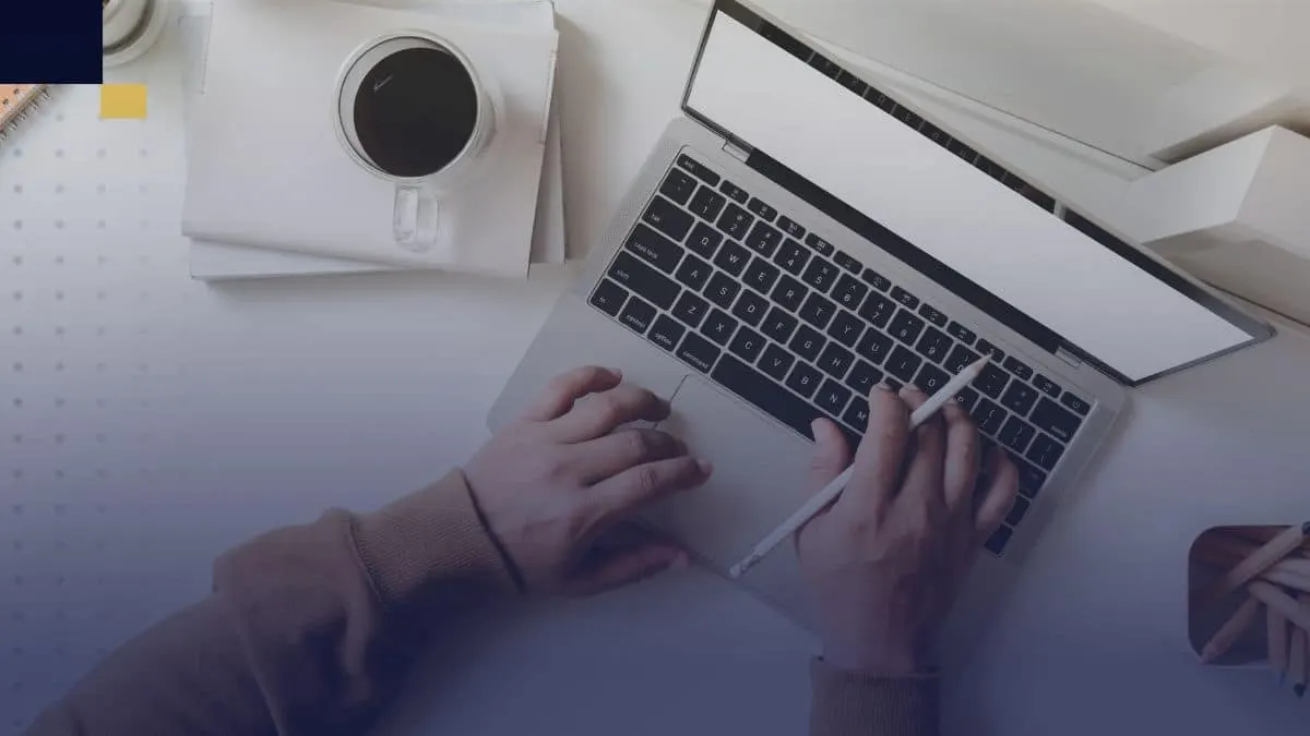 Manos escribiendo en laptop con café y cuadernos para redactar monografía académica.
