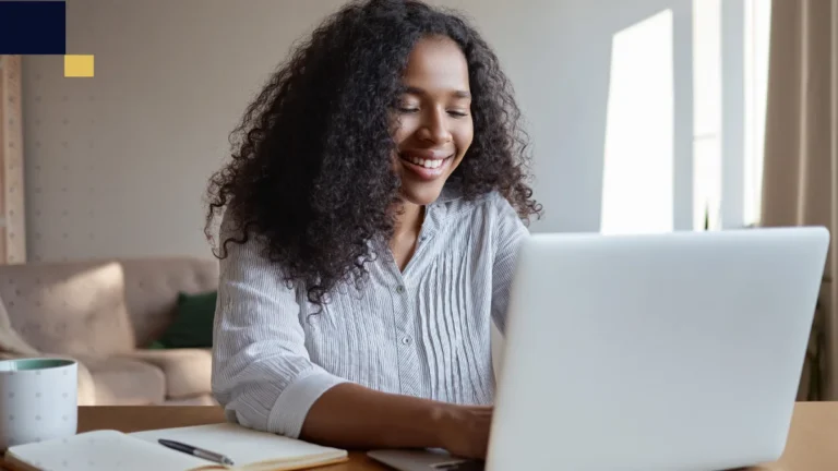 Estudiante sonriendo mientras trabaja en una laptop desde casa, imagen de Tesis y Másters Colombia sobre asesoría, corrección y apoyo académico para tesis y proyectos universitarios.