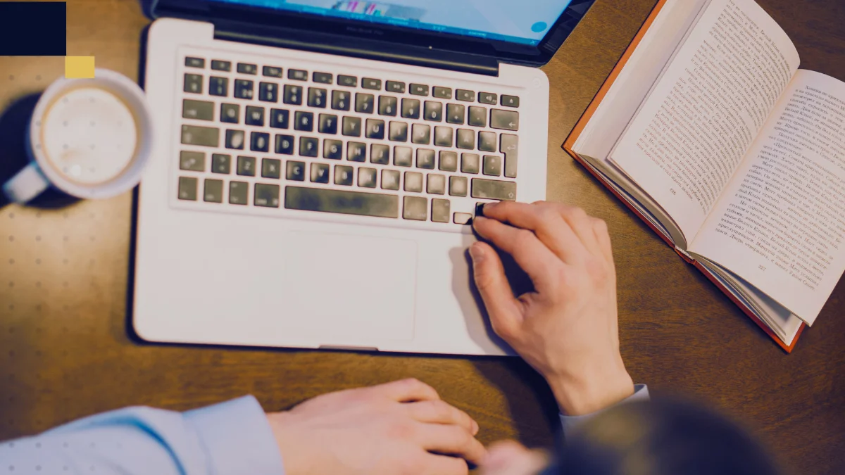 Persona trabajando en una laptop junto a un libro abierto y una taza de café, imagen de Tesis y Másters Colombia sobre asesoría, corrección y apoyo académico para tesis y proyectos universitarios.